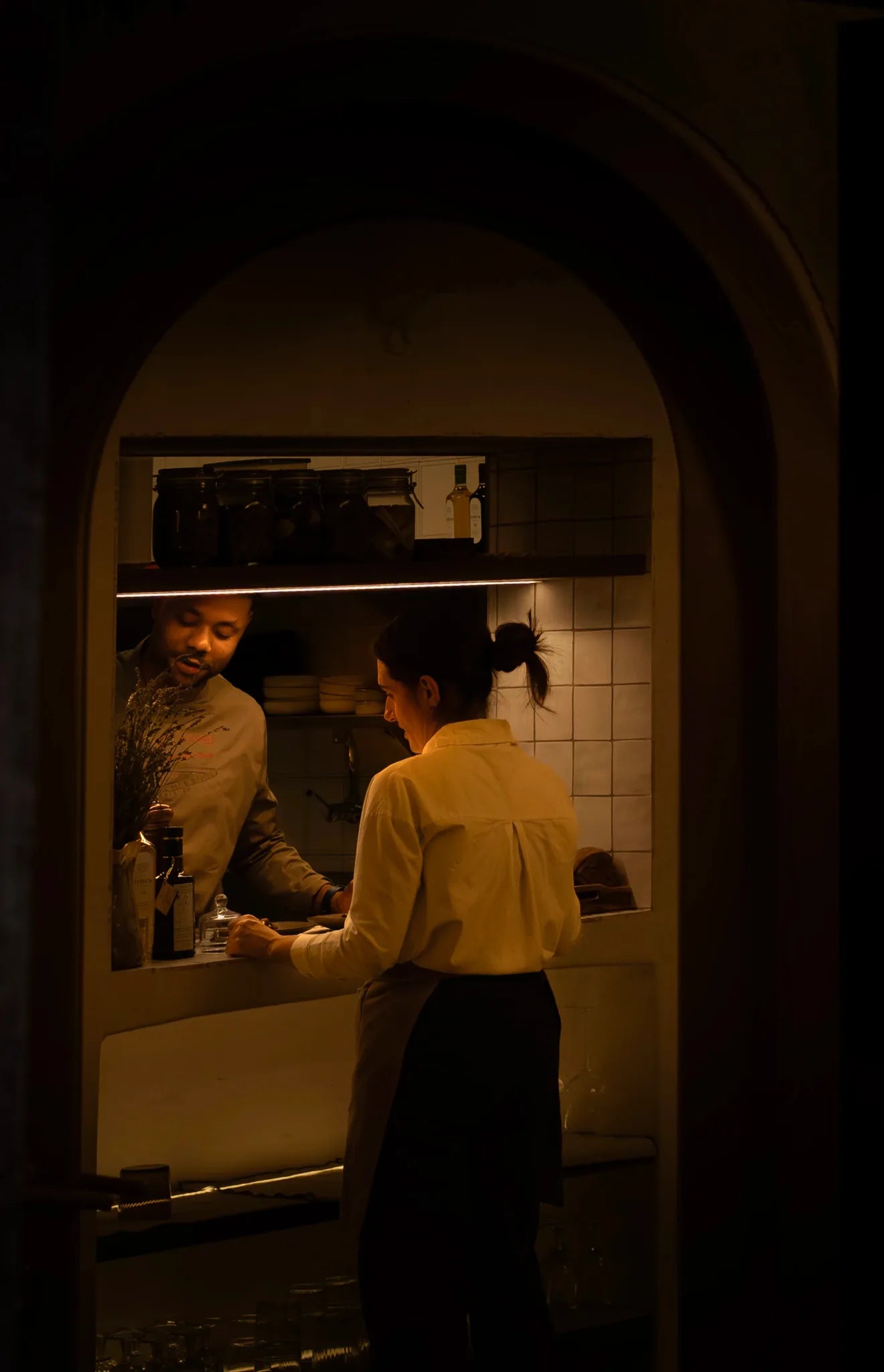 Dos personas trabajando en una cocina, vistas a través de un arco con luz cálida.