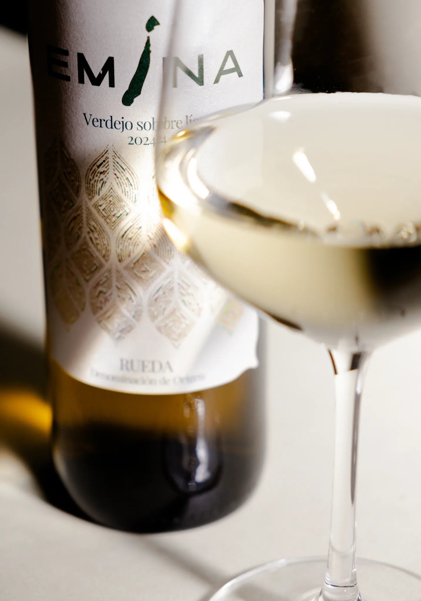 Detalle de una botella Emina y una copa de vino blanco con reflejos de luz.